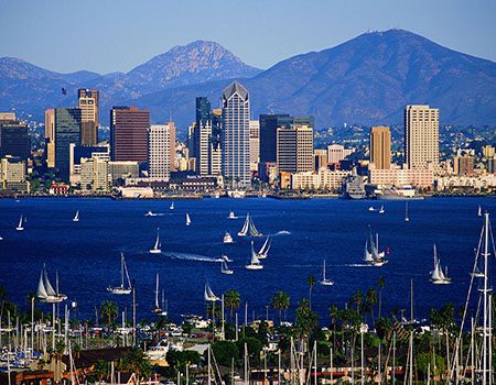 Sailboats navigating the waters in front of a bustling city skyline with mountains in the background.