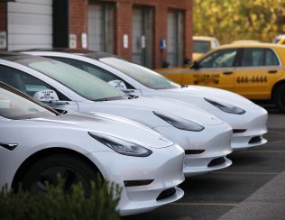 A row of white tesla vehicles parked in succession with a yellow taxi in the background.