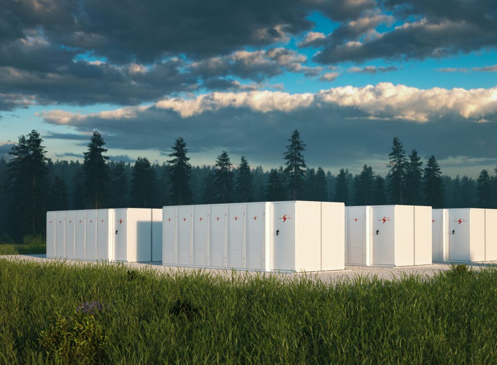 Battery energy storage system units in a field with a forest in the background and a cloudy sky above.