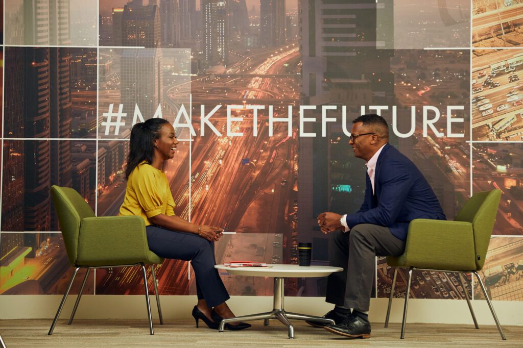Two professionals having a conversation in an office with a cityscape backdrop and a "#makethefuture" slogan on the wall.