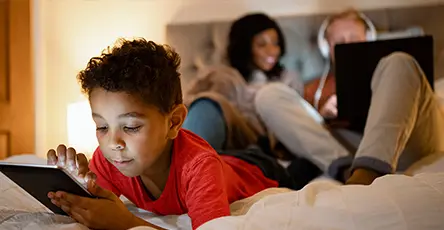 A child uses a tablet in the foreground while two adults work on a laptop in the background, all in a cozy bedroom setting.